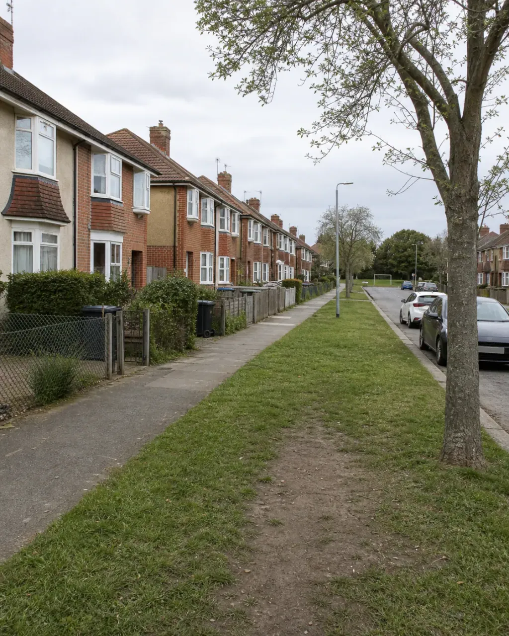 Interwar brick and render semis behind a wide grass verge on a Whitley street, a worn path crossing the green