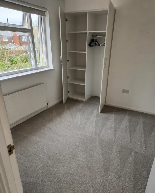 An empty bedroom mid checkout, fitted wardrobe doors open on bare shelves and hangers, carpet freshly hoovered