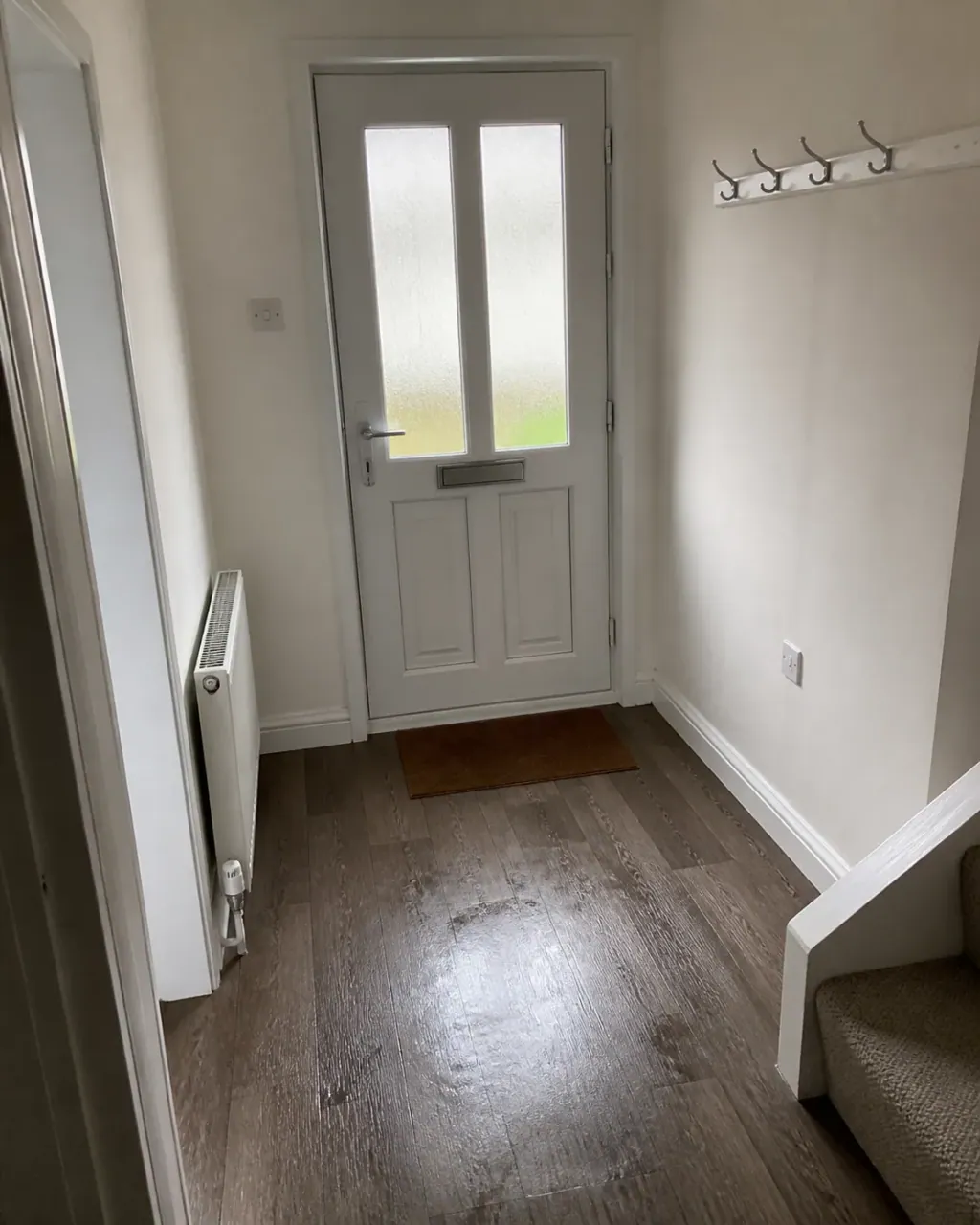 A bright hallway with a frosted glass front door, empty coat hooks and a doormat squared to the door